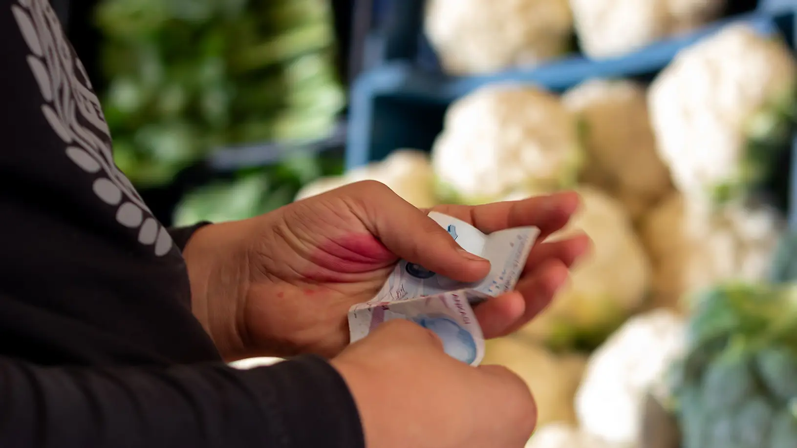 imagem com foco na mão de pessoa branca segurando dinheiro ao fundo uma pilha de couve-flor e brócolis