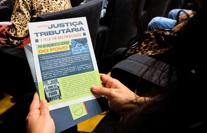 Imagem mostra as mãos de uma mulher segurando o folheto da campanha pela taxação dos super-ricos.