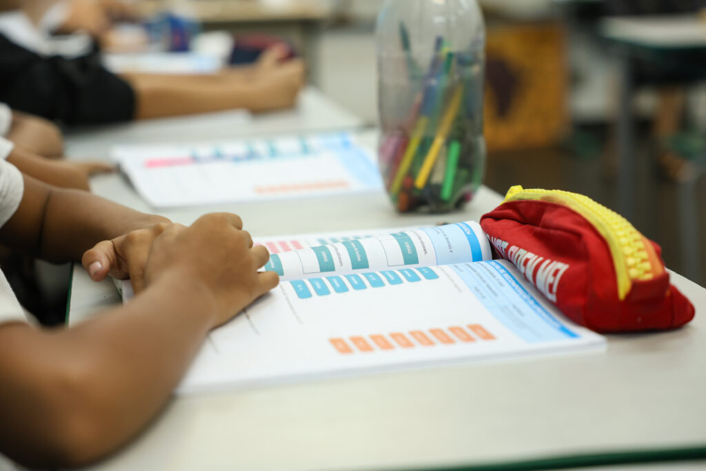 Imagem mostra os braços de uma criança sobre um livro. Ele está em uma carteira, em sala de aula. Imagem ilustra o tema da educação no relatório Inesc 2025.