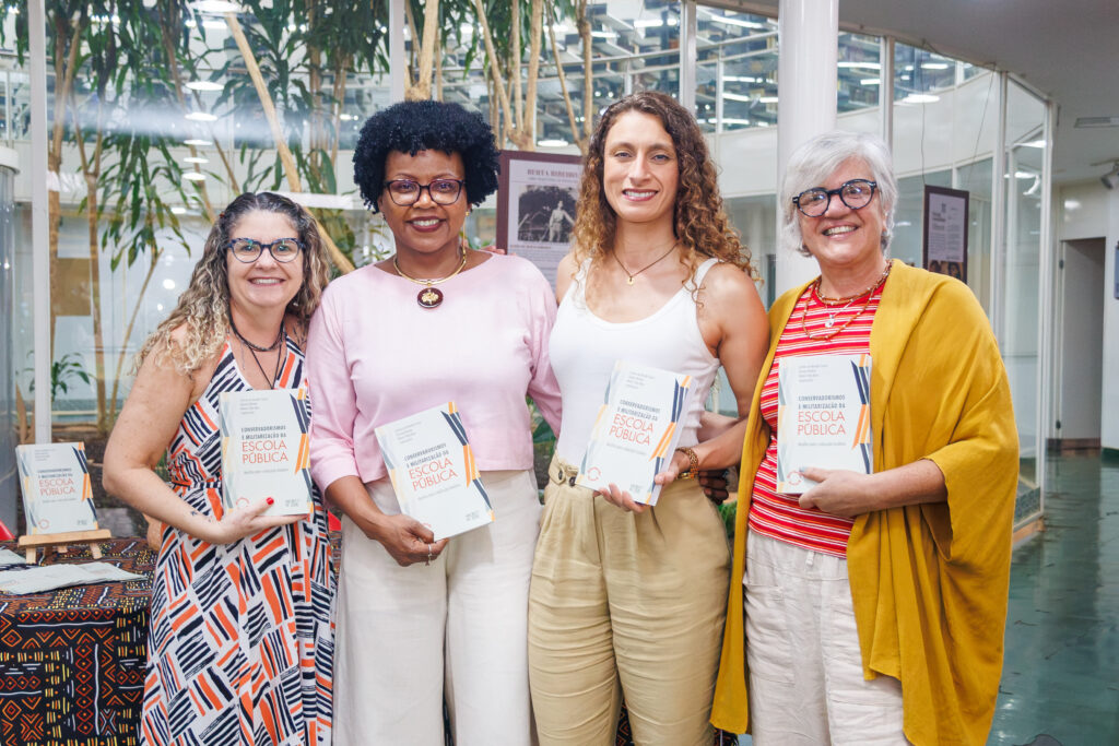 Quatro mulheres em pé, segurando o livro Conservadorismos e Militarização da Escola Pública.